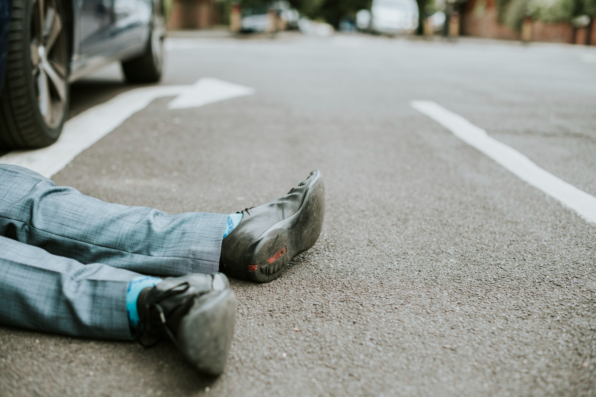 A person wearing a gray suit and black shoes, possibly a pedestrian hit by car, is lying on the asphalt of a road, with only their legs visible. A parked car and white road markings are nearby. A person wearing a gray suit and black shoes, possibly a pedestrian hit by car, is lying on the asphalt of a road, with only their legs visible. A parked car and white road markings are nearby.