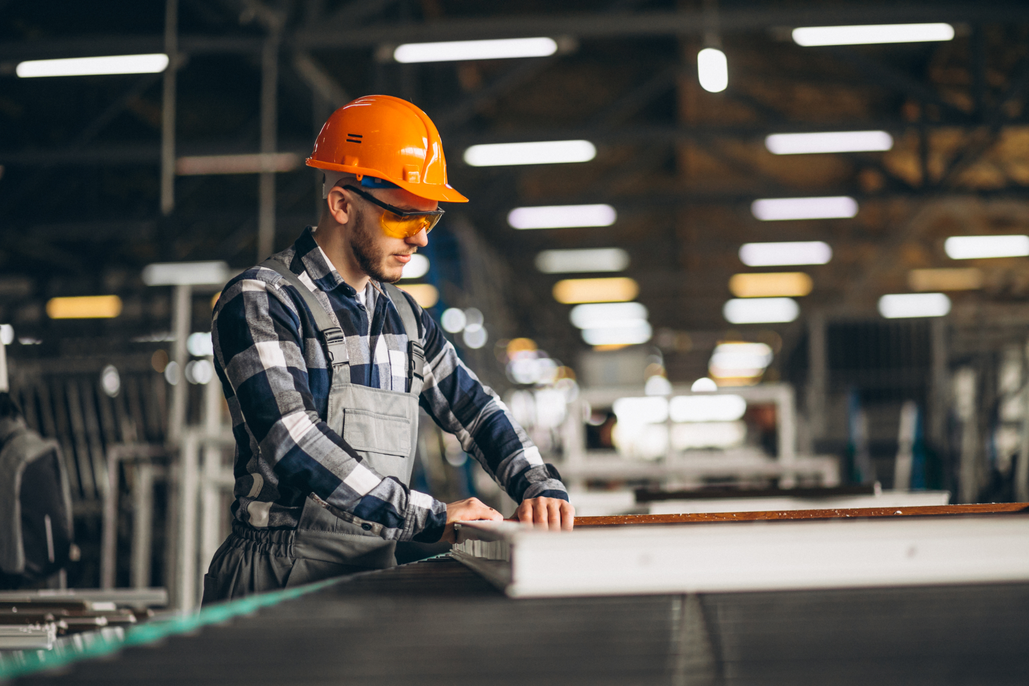 A factory worker wearing a plaid shirt, overalls, an orange hard hat, and safety glasses inspects a panel on a production line in a brightly lit industrial setting, maintaining safety to help prevent factory worker injuries. A factory worker wearing a plaid shirt, overalls, an orange hard hat, and safety glasses inspects a panel on a production line in a brightly lit industrial setting, maintaining safety to help prevent factory worker injuries.