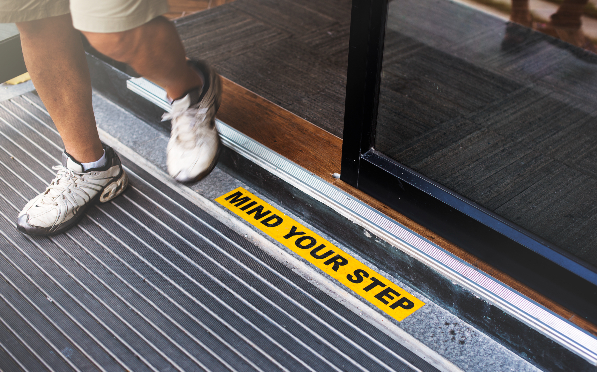 A person steps over a door threshold in a Sydney cafe, with a yellow sign on the floor that reads "MIND YOUR STEP." The sign helps prevent accidents and potential injury compensation claims.