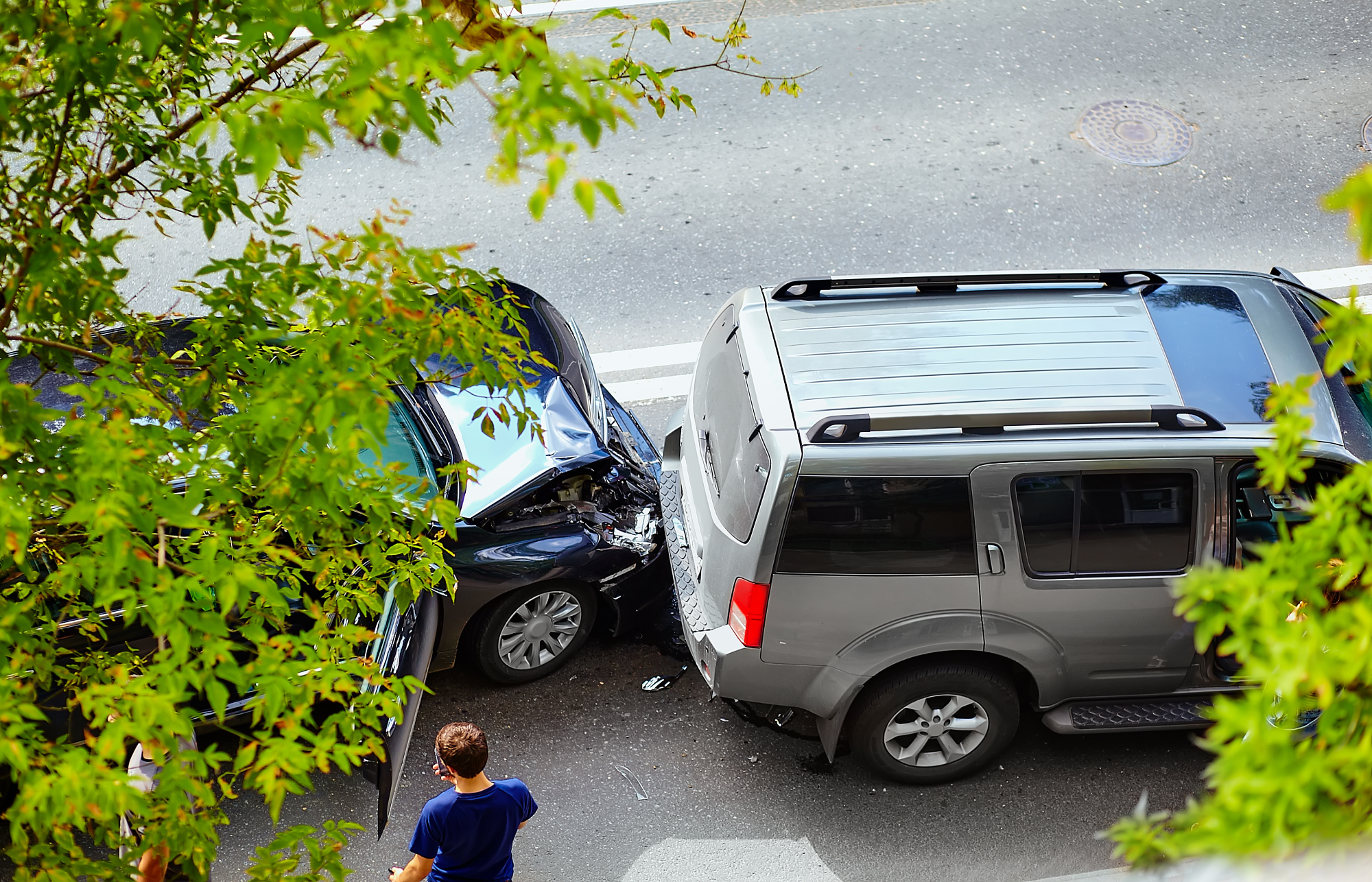Aerial view of a traffic accident shows a black car crashed into the back of a gray SUV on a city street, with a person standing nearby and green tree branches framing the image. Aerial view of a traffic accident shows a black car crashed into the back of a gray SUV on a city street, with a person standing nearby and green tree branches framing the image.