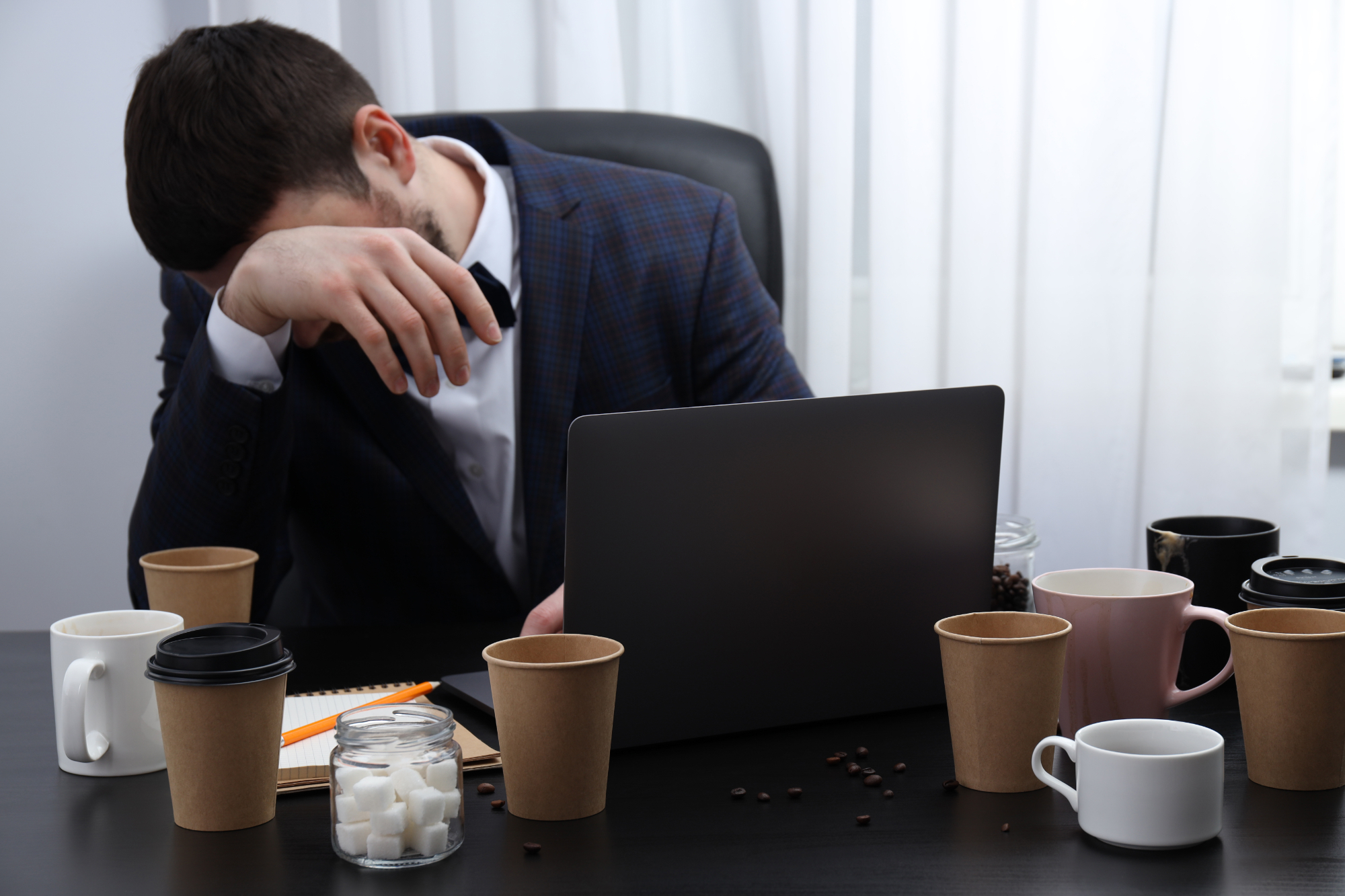 A man in a suit sits at a desk, resting his head on his hand in frustration as he faces superannuation paperwork. His laptop is open, and the desk is cluttered with empty coffee cups, a notebook, and a jar of sugar cubes.