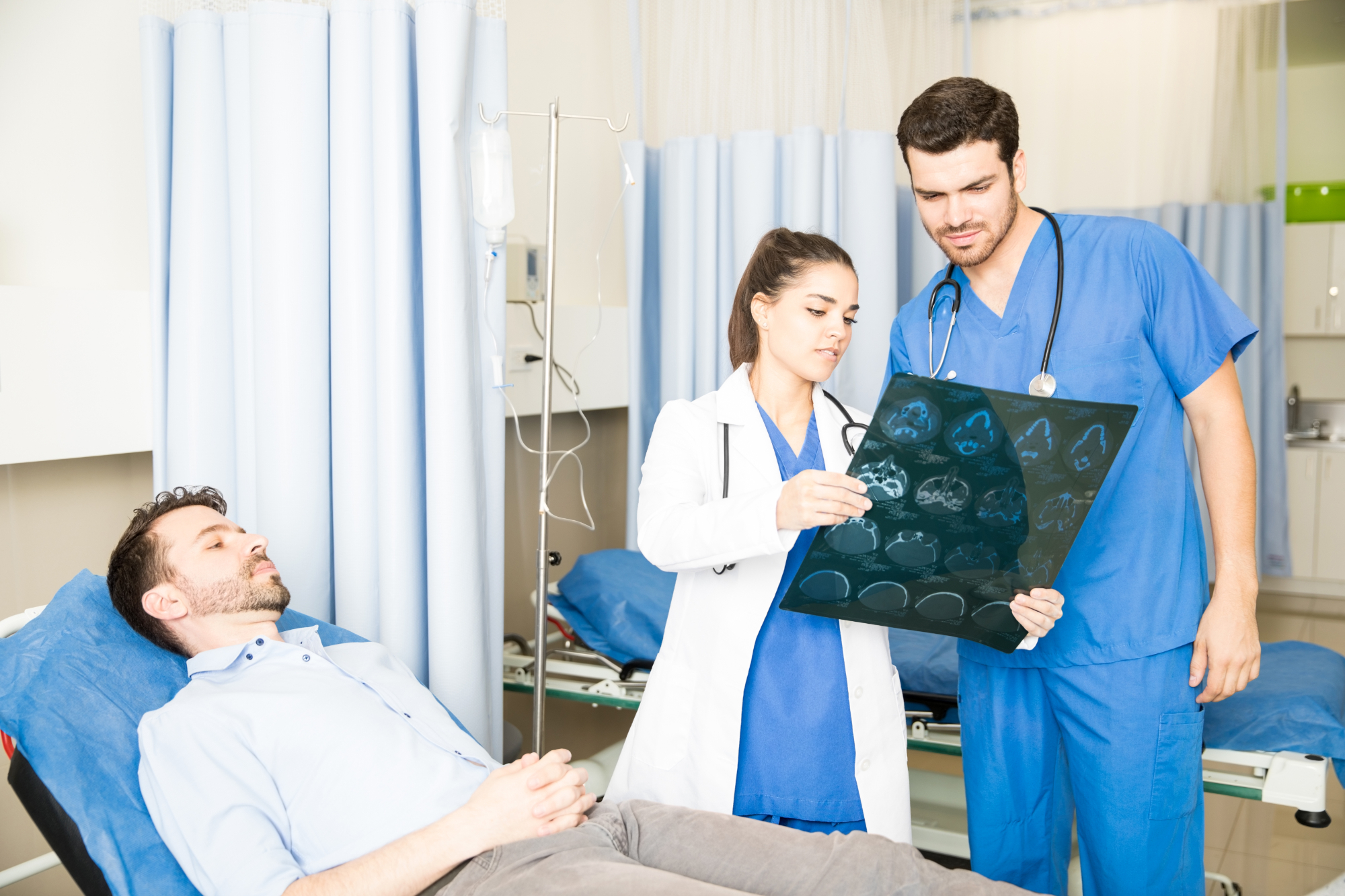 A female doctor and a male nurse examine brain scans, possibly for an Accident Compensation case, while a male patient rests on a hospital bed nearby in a medical room with blue curtains and equipment. A female doctor and a male nurse examine brain scans, possibly for an Accident Compensation case, while a male patient rests on a hospital bed nearby in a medical room with blue curtains and equipment.