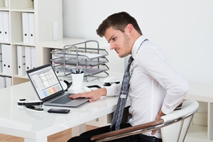 A man in business attire sits at a desk using a laptop, his strained expression and hand on his back suggesting discomfort or back pain—an issue often addressed through workers compensation. Office supplies and shelves fill the background. A man in business attire sits at a desk using a laptop, his strained expression and hand on his back suggesting discomfort or back pain—an issue often addressed through workers compensation. Office supplies and shelves fill the background.