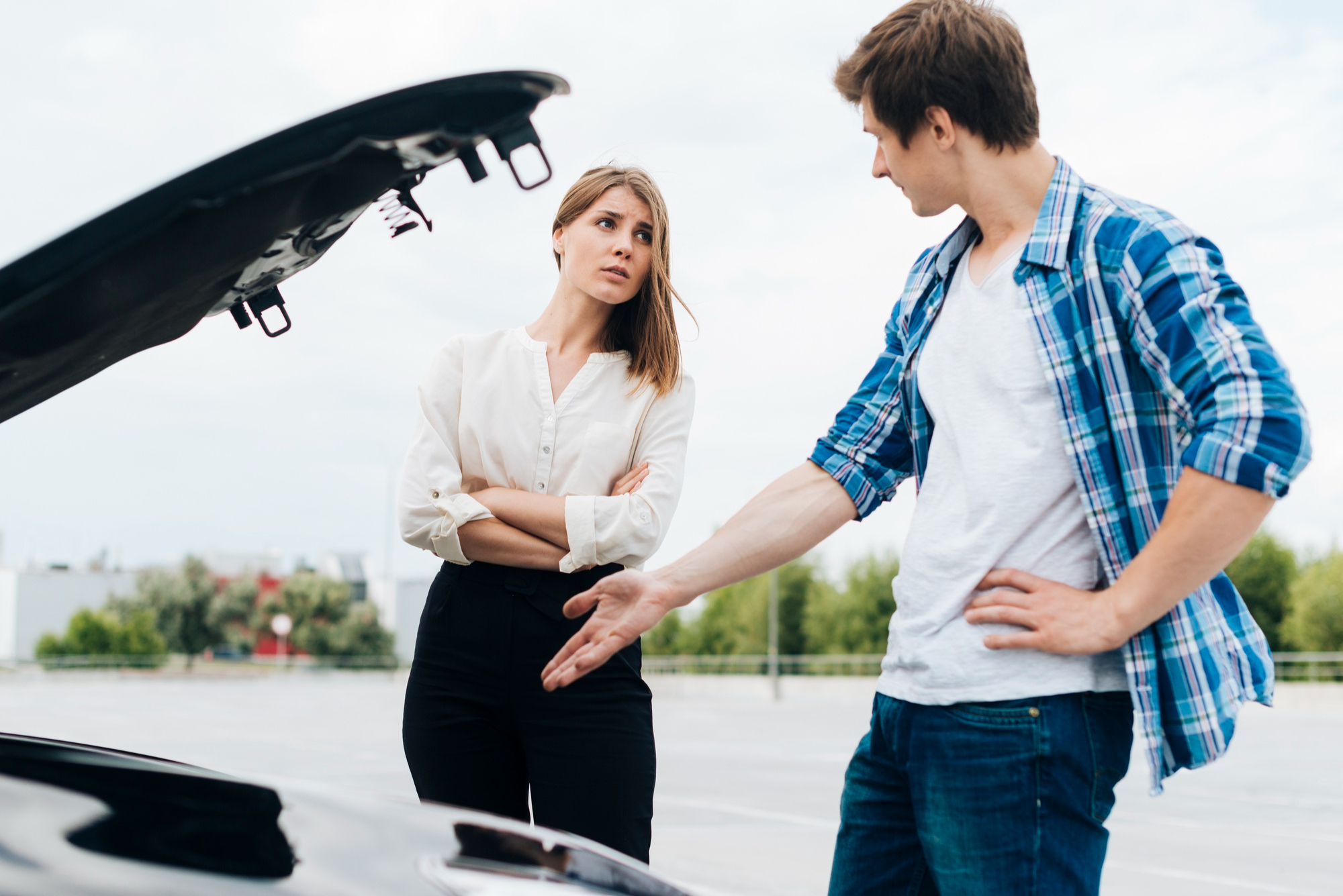 A woman stands with arms crossed beside a car with its hood open, looking at a man who gestures towards the car. They appear to be discussing motor accidents in an outdoor parking lot. A woman stands with arms crossed beside a car with its hood open, looking at a man who gestures towards the car. They appear to be discussing motor accidents in an outdoor parking lot.