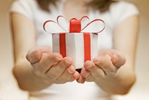 A person in a white shirt holds out a small round gift box wrapped with a red ribbon and bow, offering it with both hands—a gentle gesture contrasting the seriousness of topics like medical negligence. The background is softly blurred.