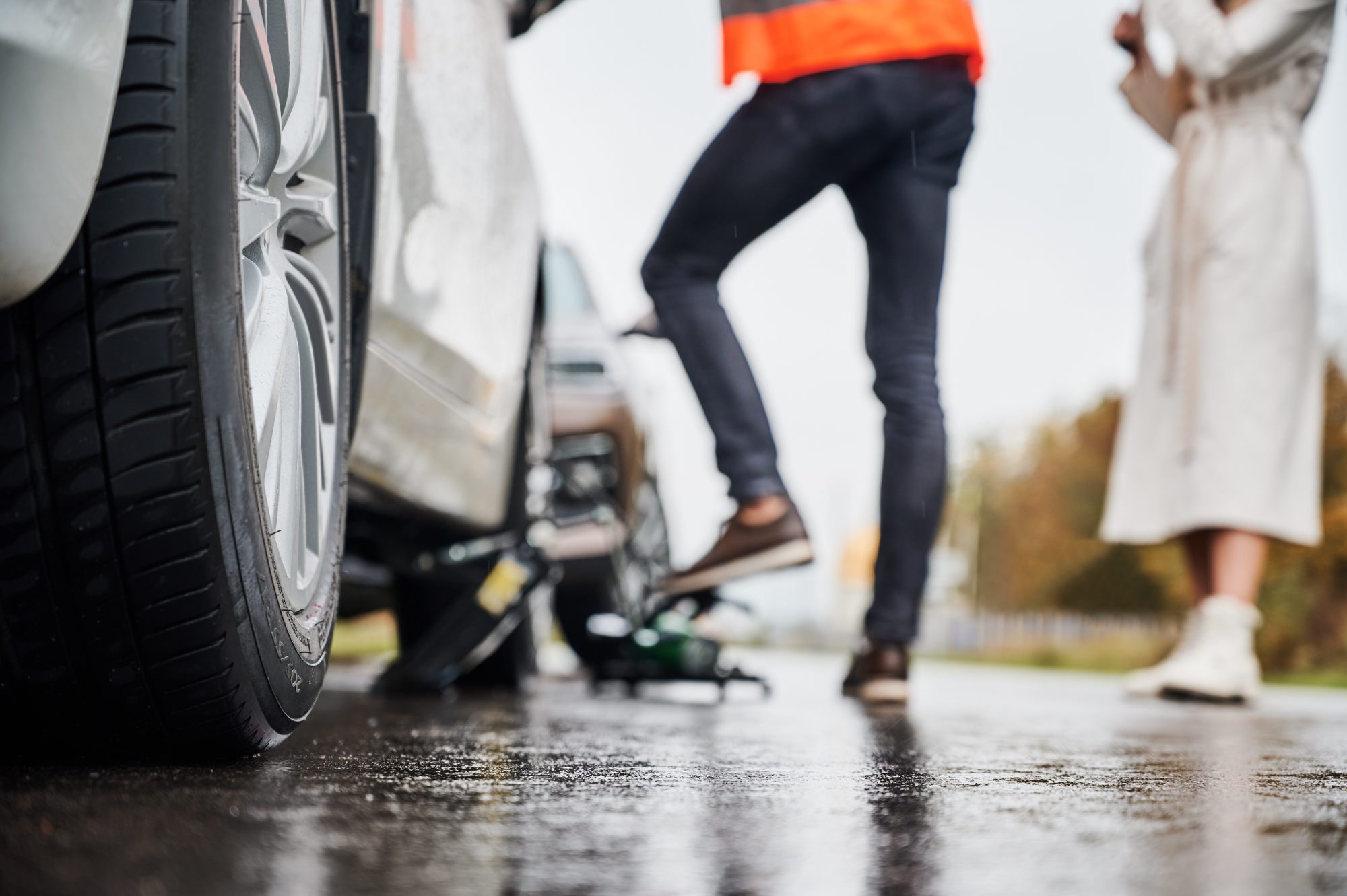 Close-up of a car tire being changed on a wet road after a car accident; one person in a reflective vest works near the car while another stands nearby in a light-colored coat. Close-up of a car tire being changed on a wet road after a car accident; one person in a reflective vest works near the car while another stands nearby in a light-colored coat.
